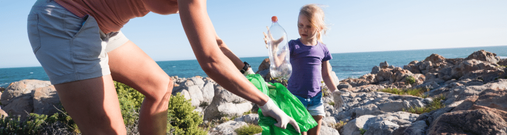 Woman holding plastic bag open with young girl putting plastic bottle into it, by the ocean