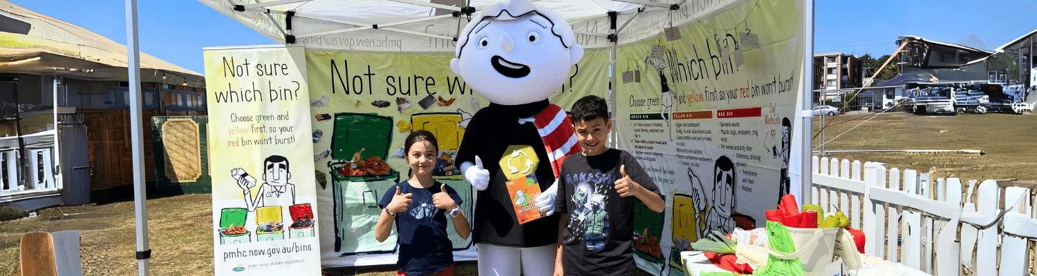 Black and white large cartoon character mascot standing next to two children giving a thumbs up, at a market stall for waste education.